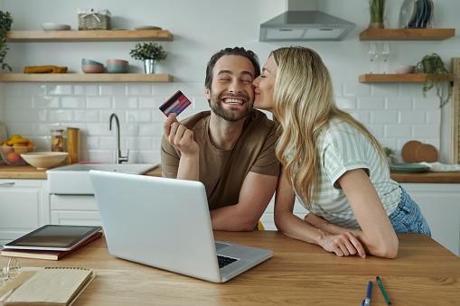 Cheerful young couple using credit card while shopping online from domestic kitchen