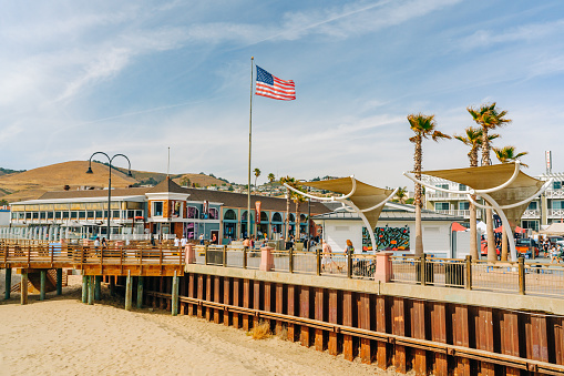 Pismo Beach, California, USA - June 3, 2022. Wooden boardwalk along the shore, wide sandy beach, and plaza in downtown of Pismo Beach city, California central coast