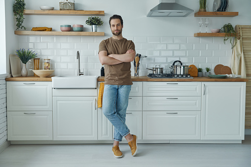 Full length of handsome young man leaning at the kitchen desk and looking at camera