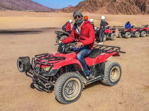 Tourist man dressed in a kufiya sits behind the wheel of the quad bike in the Sinai desert against the backdrop of a caravan of ATVs, Egypt. Active entertainment for tourists in Sharm El Sheikh