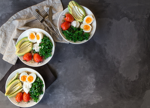 Quinoa buddha bowls with baked tomatoes, avocado, kale, boiled eggs and greek yogurt on gray concrete background. Healthy breakfast. Top view. Copy space area.