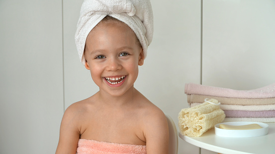 Portrait of little girl looks at camera and laughs in bathroom. Happy child after bathing with wrapped towel on head. Baby care.