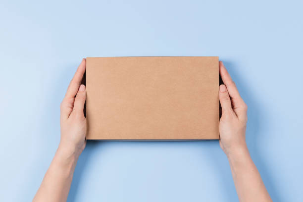 istock Top view to female hands holding brown cardboard box on light blue background. Mockup parcel box. Packaging, shopping, delivery concept 1402351231