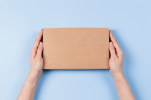 Top view to female hands holding brown cardboard box on light blue background. Mockup parcel box. Packaging, shopping, delivery concept.