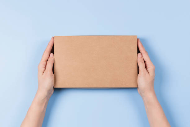 Top view to female hands holding brown cardboard box on light blue background. Mockup parcel box. Packaging, shopping, delivery concept stock photo