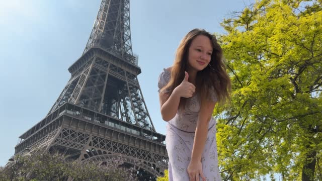 beautiful young teenage girl in paris on the background of the eiffel tower in a long chic dress smiles and shows the class looks at the camera she has long blonde hair and around good weather behind her bright green tree can be used to advertise trips to