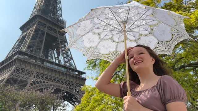 beautiful young teenager girl in paris on the background of the eiffel tower in a long elegant dress in the style of romanticism walks with an umbrella from the sun and smiles at her long blonde hair and around good weather and in the background the eiffe
