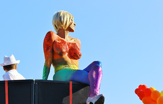 Transsexual woman covered in rainbow body paint and glitter. Sitting on a speaker-system. Gay Pride. Clear sky.