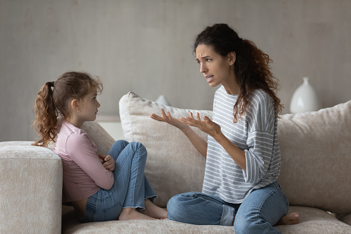 Annoyed emotional strict mother lecturing offended little daughter for bad behavior, sitting on couch at home, unhappy serious mom scolding shouting upset child, family generation conflict concept