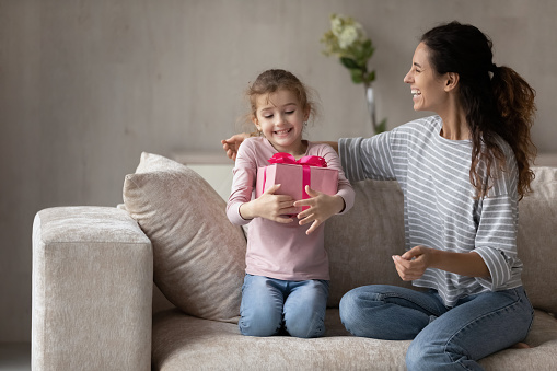 Loving caring happy mother congratulating little daughter, presenting pink gift box, sitting on cozy couch, young family excited smiling mom with adorable girl kid celebrating birthday at home