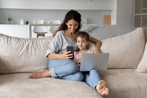 Happy mother with cute daughter having fun with electronic devices at home, using smartphone and laptop together, smiling mom and adorable girl kid chatting online by video call, watching cartoons