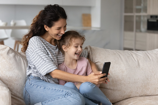 Overjoyed mother with cute girl daughter having fun with smartphone, looking at screen, chatting online by video call, happy mom and adorable kid taking selfie, spending leisure time with device