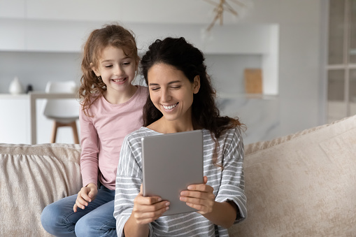 Close up happy mother with adorable daughter using tablet together, looking at screen, smiling young mom and cute girl kid chatting online by video call, having fun with electronic  device at home