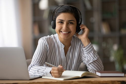 Portrait of smiling beautiful millennial Indian woman wearing headphones with microphone, writing notes in copybook listening interesting educational online lecture webinar, e-learning concept.