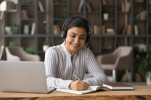 Happy beautiful Indian ethnicity woman in wireless headset with mic writing notes holding video call meeting or watching educational webinar lecture, improving knowledge on online courses.