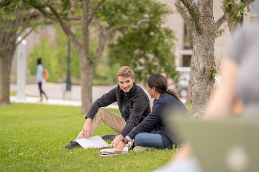 Two University students sit outside on the grass together as they study.  They are each dressed casually and have a textbook open in front of them as they study under a tree on campus.