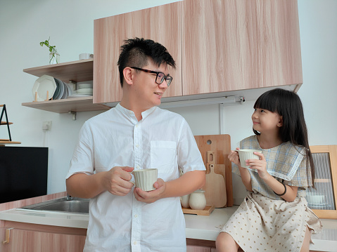 Parenthood. Family. Love. Asian Chinese father and his little daughter are holding a cups of drink in the kitchen at home.