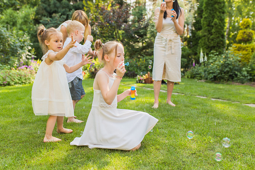 Blow and catch soap bubbles. Girl in white dress at backyard. Parents and children. Outdoor recreation. Childhood. Leisure activity. Summer picnic vacation. Rest. Brothers and sisters. House rental