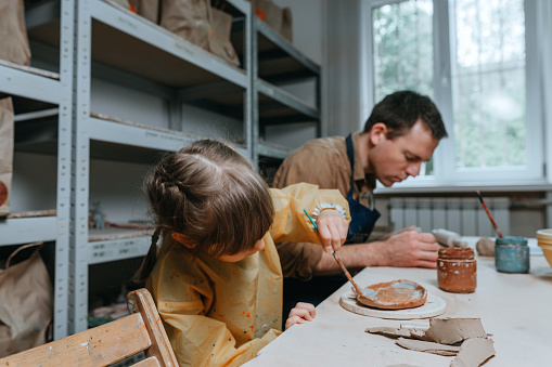 Father and his daughter at a master class of pottery clay modeling in the workshop. Young man and little girl working with clay. Happy family leisure. Idea for DIY family activity.