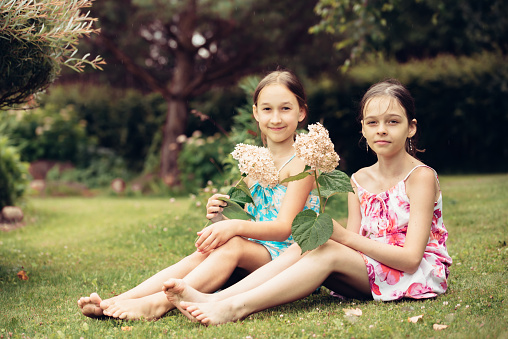 Two little barefoot girls in light summer dresses pose sitting on the ground in a summer park. Children with flowers in their hands are resting in the village, summer holidays. Carefree childhood.