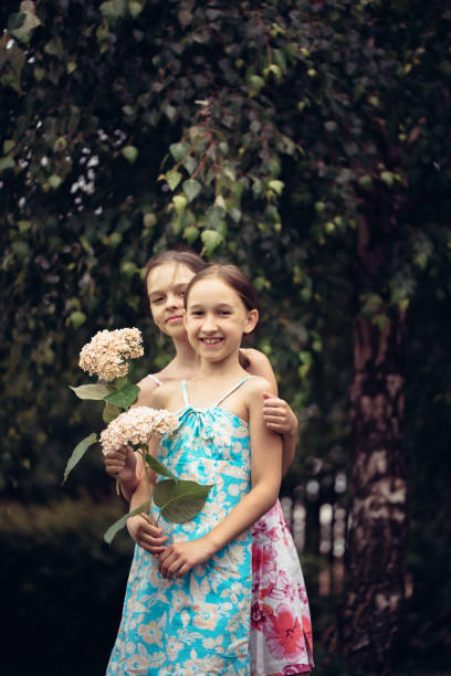 Two little girls in light summer dresses are walking in the garden on a warm summer day. Children with flowers in their hands are resting in the village, summer holidays. Carefree childhood.