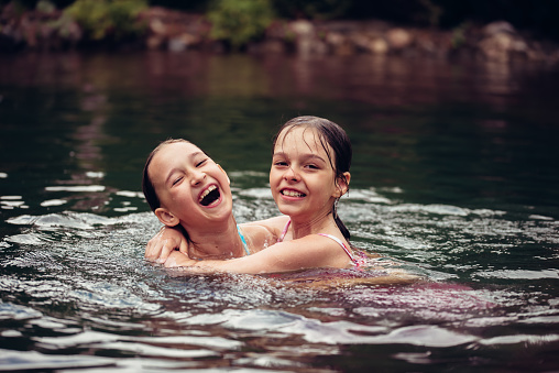 Two little girls in summer dresses swim in a pond outdoors. Children rest in the village, summer vacation. Carefree childhood. The concept of relaxation and fun. Children swim in the pool.