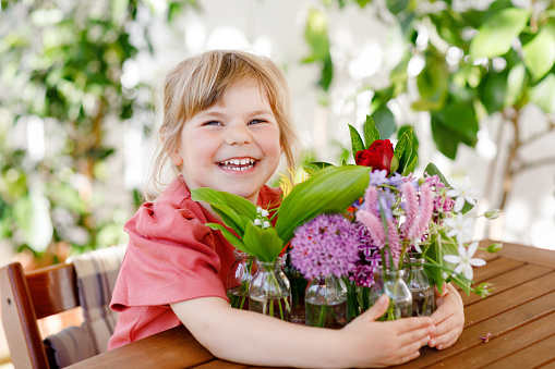 Little preschool girl with flower bouquet at home. Toddler child putting colorful garden summer flowers in small bottles with water. Home activities for kids. Flowers in rainbow colors. Happy child