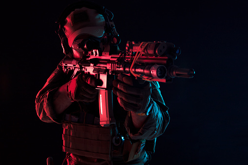 Photo of a fully equipped soldier in armored vest, helmet, face glasses and protection kneeling and attacking with rifle on dark background