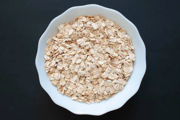 Oat flakes, top view of organic and vegan cereal in white cup. Popular healthy oat meal isolated on dark background, selective focus.