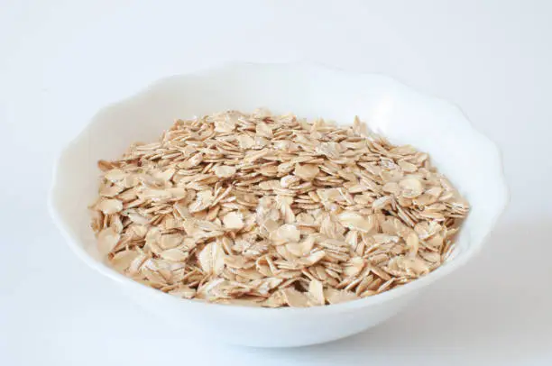 Oat flakes, angled profile view of natural healthy oat flakes in white bowl for morning breakfast. Vegan food isolated on white background, selective focus.