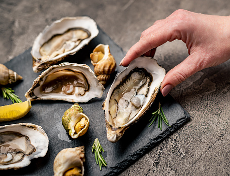 Woman hand holding fresh oyster taken from black platter with cooked snails, seafood and lemon