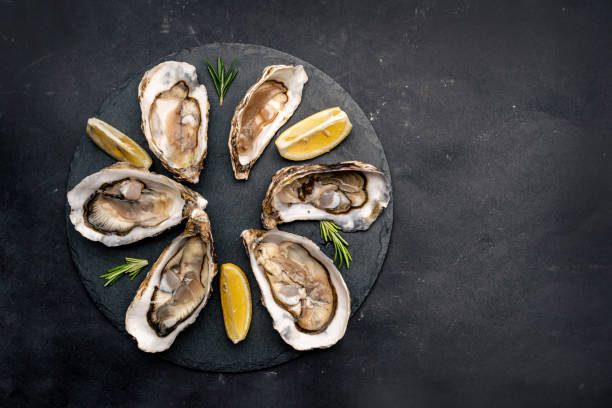 Oysters with lemon on platter Raw seafood oysters served with lemon on round black plate isolated on black background oyster stock pictures, royalty-free photos & images