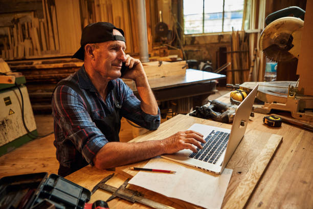 istock Senior carpenter works on the computer and talks phone with client in the workshop 1402451445