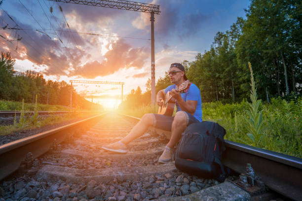 Handsome guy who was late on train sitting on rail and play acoustic guitar. Traveling and lifestyle concept.