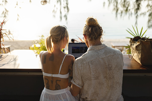Rear-view of unrecognizable modern young Caucasian couple working on laptop from coffee shop, on their vacation at Thailand