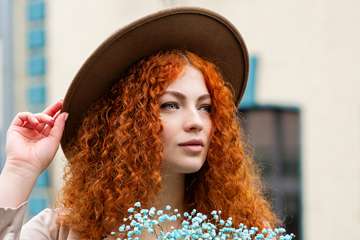 Curly Red-haired Girl in Felt Hat, Raincoat Looks Away. Young Woman Holds Bouquet Blue Flowers on City Street in Spring Cloudy Rainy Evening. Close Up Portrait