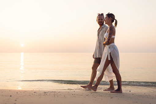 Side view of carefree Caucasian couple holding hands while walking on the beach during sunset