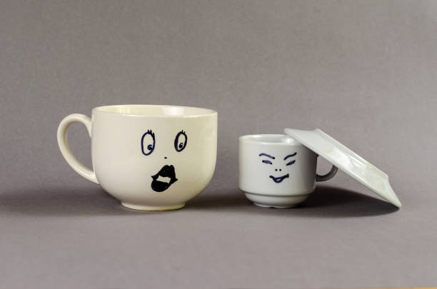 istock Two ceramic cups against a gray background. A large cup with a drawn surprised woman's face and puffy lips. A small cup with a man's face and a saucer imitating a hat removed in front of the lady. 1402481089