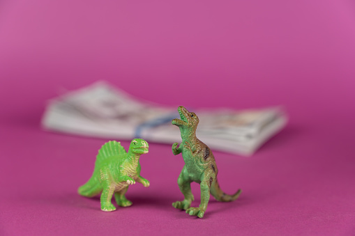 Dinosaur miniatures and a wad of bills against a purple background. A wad of cash in the background . Green figures of animals of prey standing on their hind legs. Selective focus.