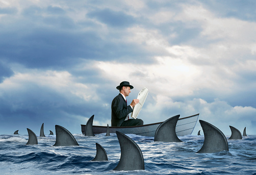 A man ignores the danger that surrounds him as he nonchalantly reads a newspaper  in a small boat that is stranded at sea and is surrounded by a swarming school of sharks.