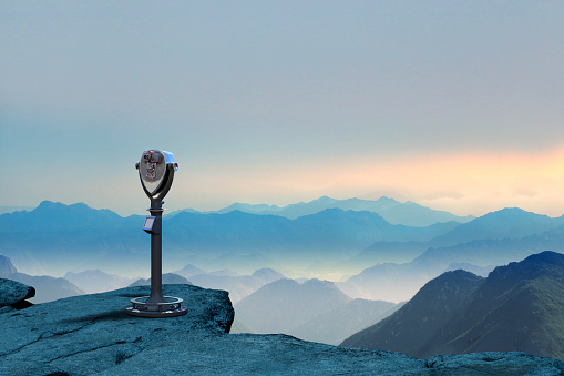 A telescope stands at the edge of a cliff as it looks out toward a dramatic landscape of overlapping mountain ridges during a late afternoon.