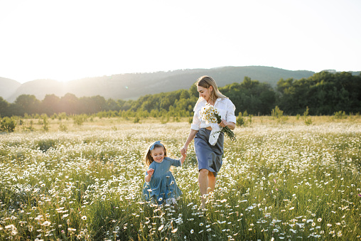 Cute beautiful young woman holding hands and play with little child girl walk in chamomile field with blooming flowers over nature background. Family lifestyle concept. Mom and baby daughter in grass