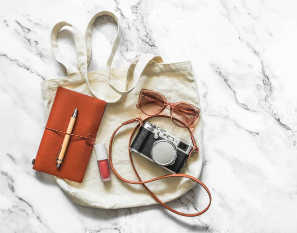 istock Women's eco textile shopper with a camera, sunglasses, leather notebook on a light marble background, top view 1402500429