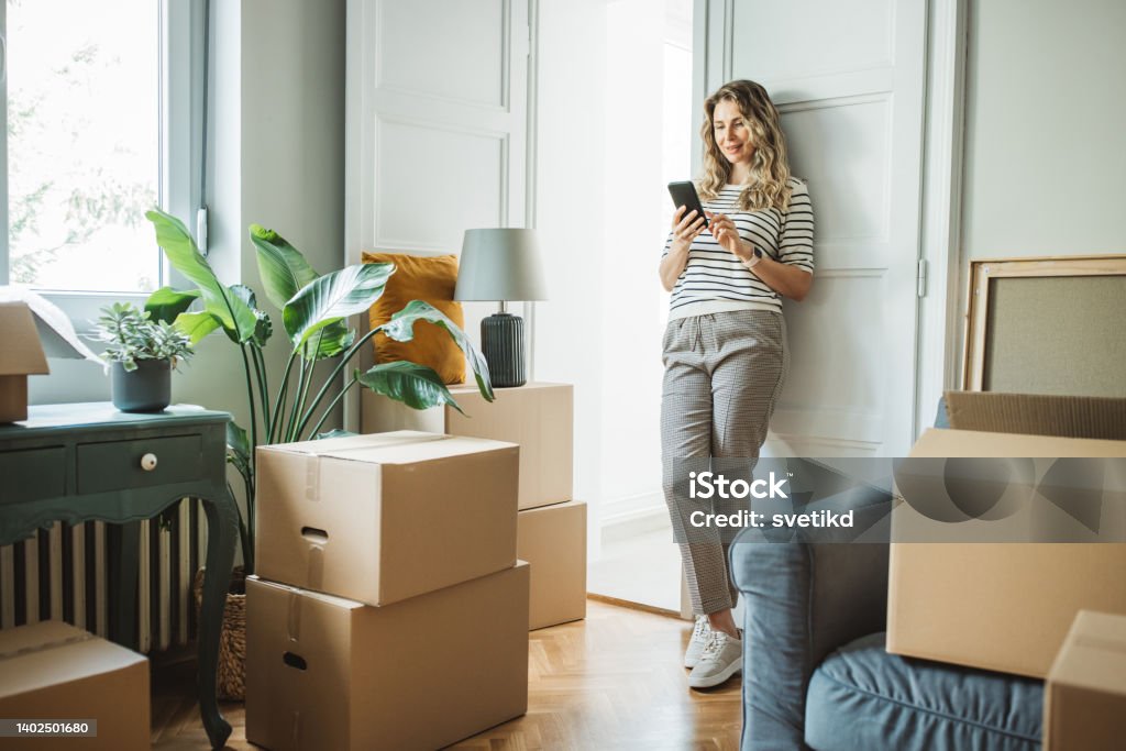 Mature woman with Moving Boxes in New Home Mature woman moves in to new home, unpacking boxes and enjoying in her new home. She is resting and using smart phone. Relocation Stock Photo