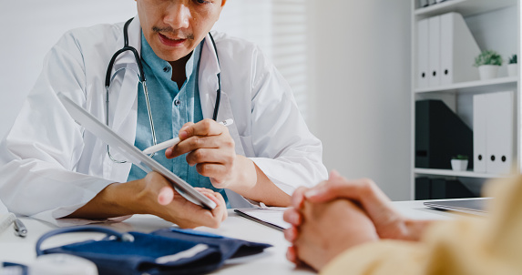 Close up of mature Asian man doctors medical results during consultation on tablet computer with woman patient in health clinic. Medical health care concept.