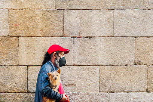 Senior man wearing red cap and face mask, walking, holding small dog. Stone wall in the background. Lugo city, Galicia, Spain.