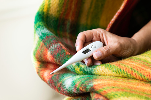 Unrecognizable female hand in multicolored blanket is holding thermometer in hand and is checking her fever.