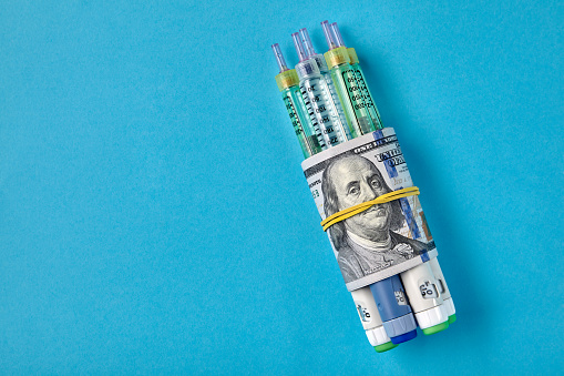 Insulin syringe pens wrapped in dollar bills on a blue background. Pharmaceutical monopolists raise drug prices. Top view with copy space