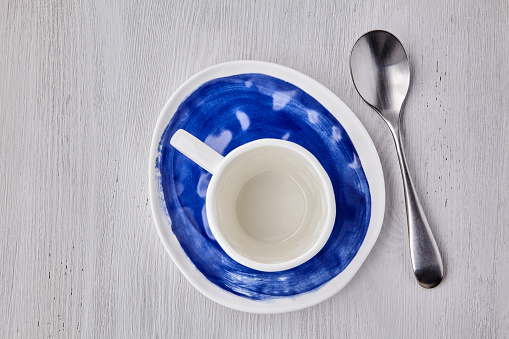 Empty white cup for hot drinks with a spoon on a blue plate on a white wooden table. Top view menu with copy space. Flat lay