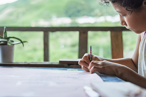 brown-skinned, low-income latina student doing her school homework at home. girl drawing a picture on a large pink poster with her colored pencil in her right hand. concept of poverty